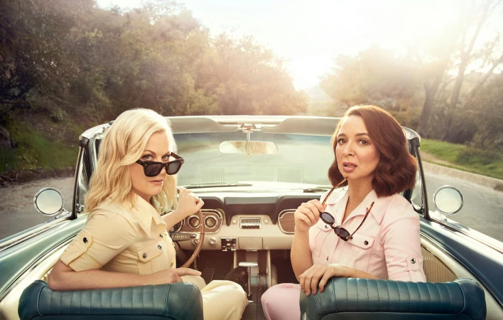 Two women seated in a vintage convertible car with bright sunlight filtering through trees in the background