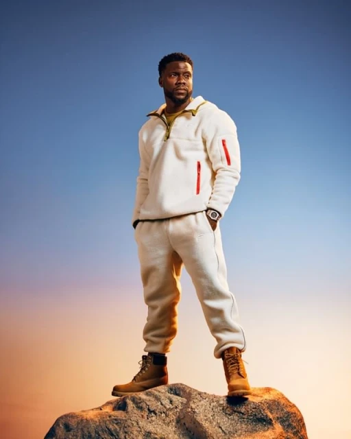 Eye-level shot of Kevin Hart posing on a rock formation against a gradient sky, wearing a white fleece tracksuit and brown boots.