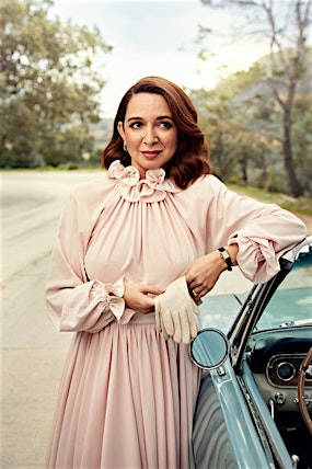 Portrait of a brunette woman in a pink dress leaning on a blue vintage car on a tree-lined road, looking to the right.