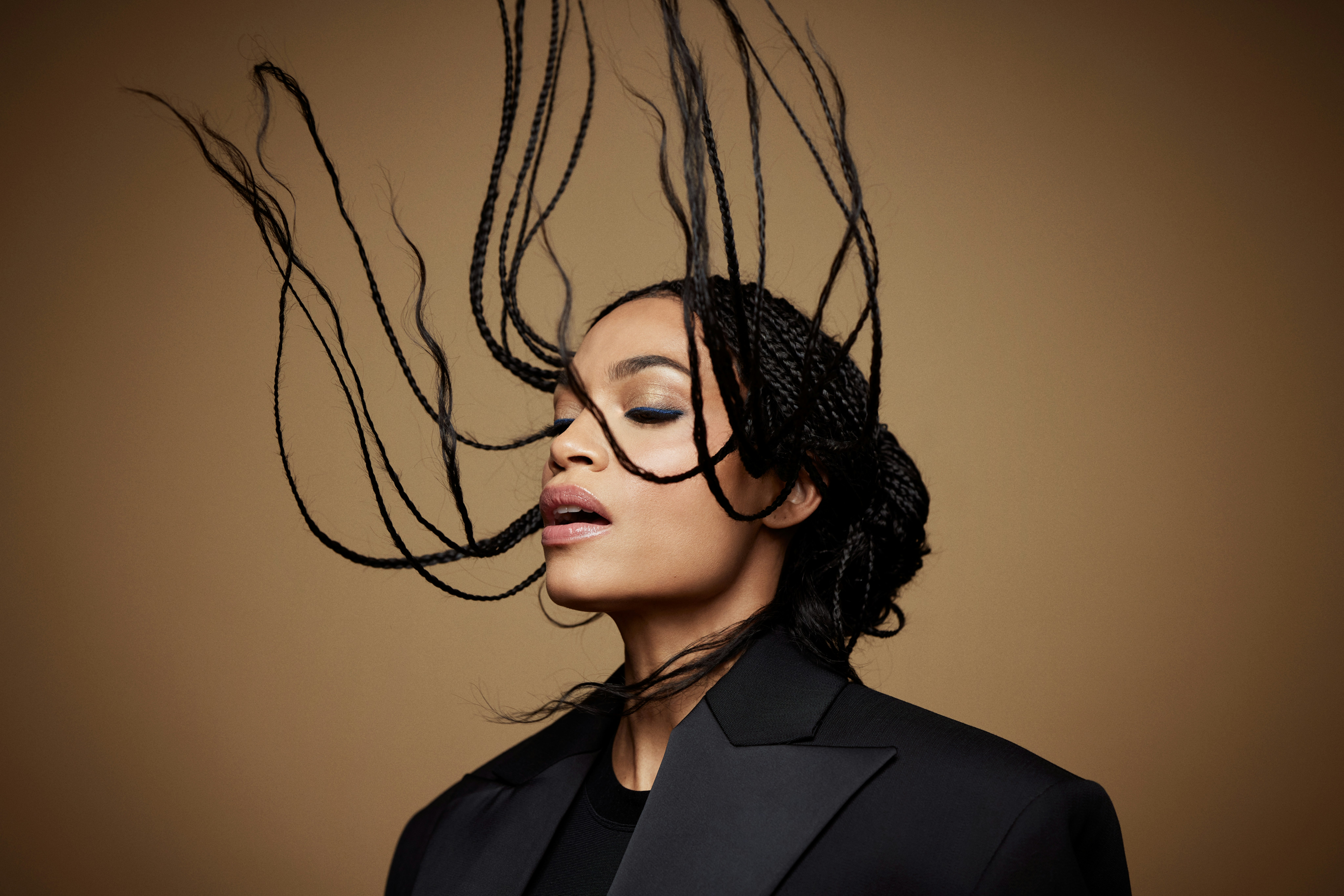 Portrait of a Black woman in a black blazer with her braided hair tossed upward against a warm brown background