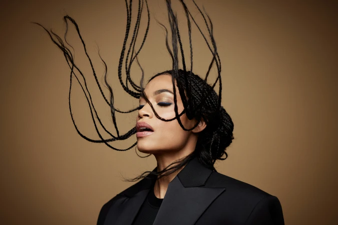 Portrait of a Black woman in a black blazer with her braided hair tossed upward against a warm brown background