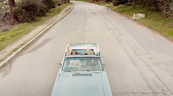 Two women enjoy a sunny drive in a vintage convertible on a winding road surrounded by lush greenery, captured from an elevated perspective.