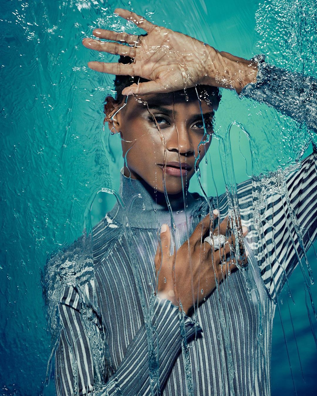 Striking portrait of a Black woman with water cascading over her face and hands against a teal background, showcasing a dramatic and captivating image.