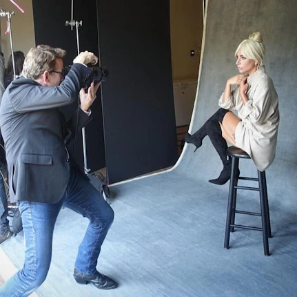 Blonde woman poses on stool in photo studio as a photographer in a suit and jeans shoots with professional camera and lighting gear.