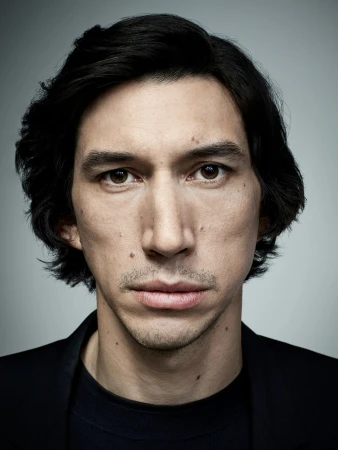 Close-up studio portrait of actor Adam Driver looking directly at the camera against a neutral grey background.