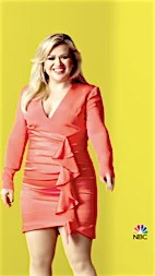 Kelly Clarkson posing in a coral dress with ruffles against a yellow background in a vertical portrait, smiling for the camera.
