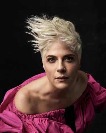 Portrait of Selma Blair with short platinum blonde spiked hair wearing a hot pink off-the-shoulder dress against a dark background