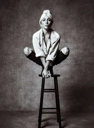 Lady Gaga crouching on a wooden stool in a white shirt and black boots against a textured background by Art Streiber