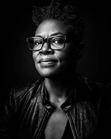 Black and white portrait of a black woman wearing black-rimmed eyeglasses and a leather jacket against a dark background.