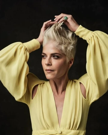 Portrait of Selma Blair with short blonde hair wearing a yellow dress holding her hands to her hair by Art Streiber