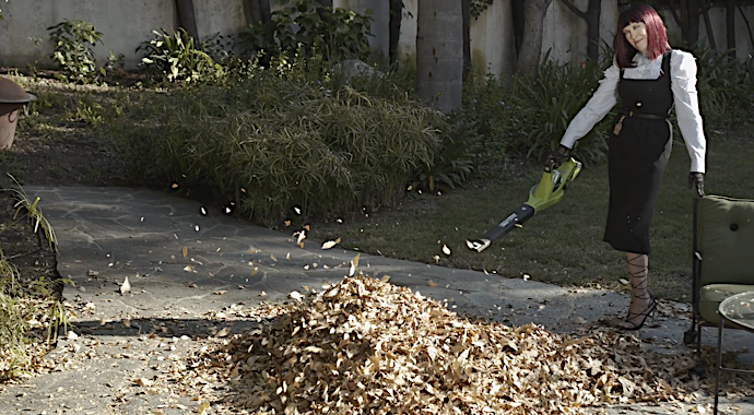 Woman in a black dress and white shirt using a leaf blower to move a pile of leaves on a concrete driveway in a yard.