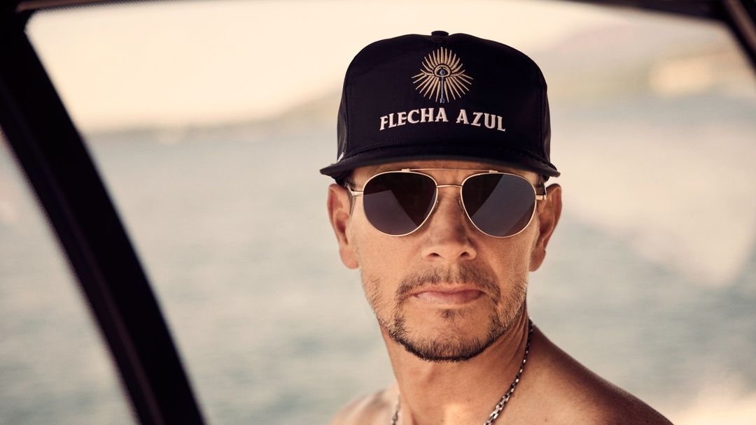 Close-up of a mature adult man wearing sunglasses and a black baseball cap that has 'FLECHA AZUL' text on it with a sun graphic.