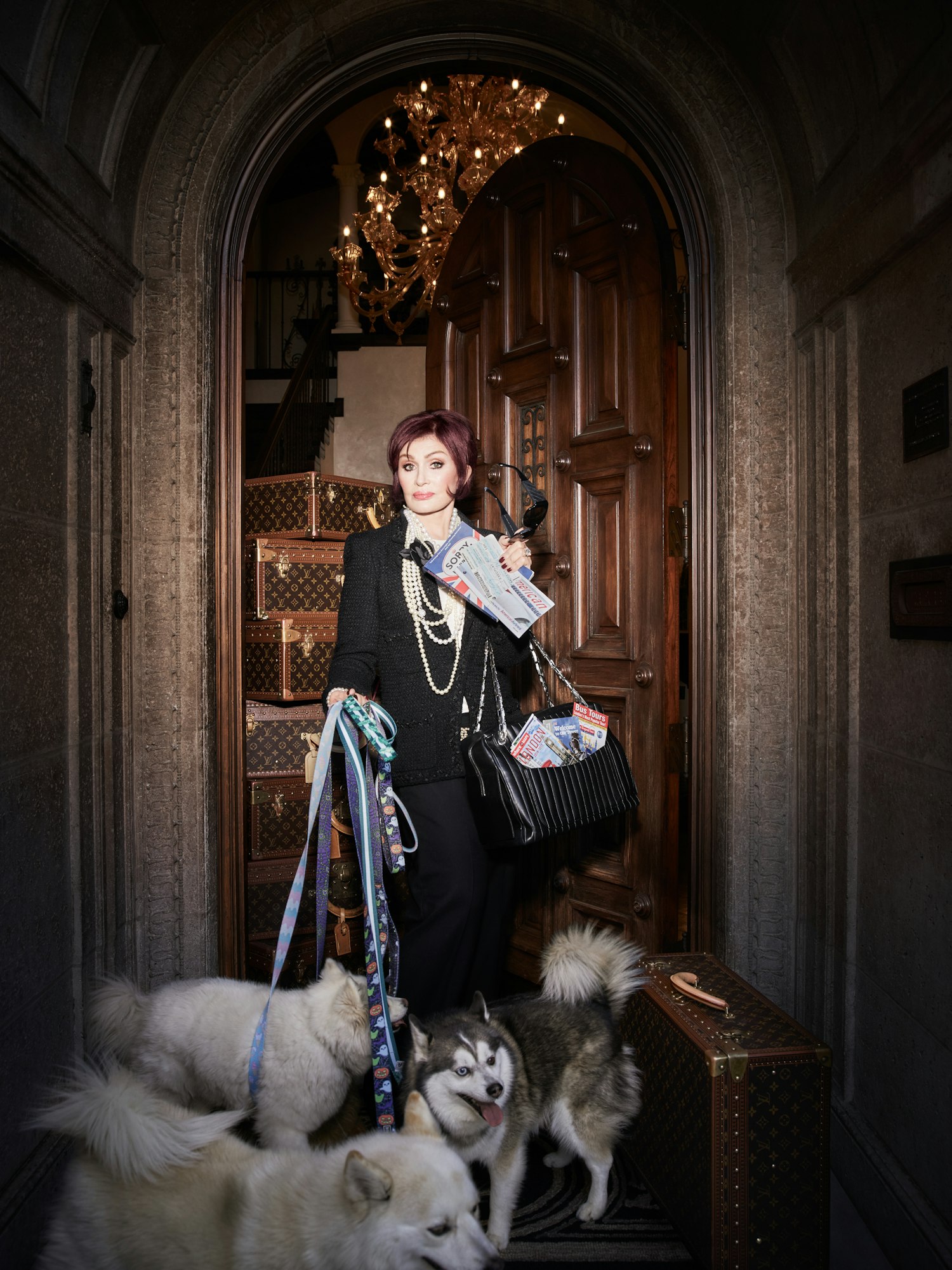 Sharon Osbourne poses with dogs and Louis Vuitton luggage in a grand doorway, wearing a black pantsuit and pearl necklace.