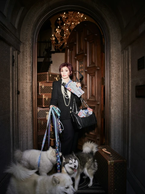 Sharon Osbourne poses with dogs and Louis Vuitton luggage in a grand doorway, wearing a black pantsuit and pearl necklace.
