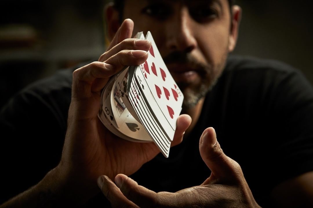 An adult man with a beard showcases a flourish with a deck of playing cards in his hands in an indoor dark setting.