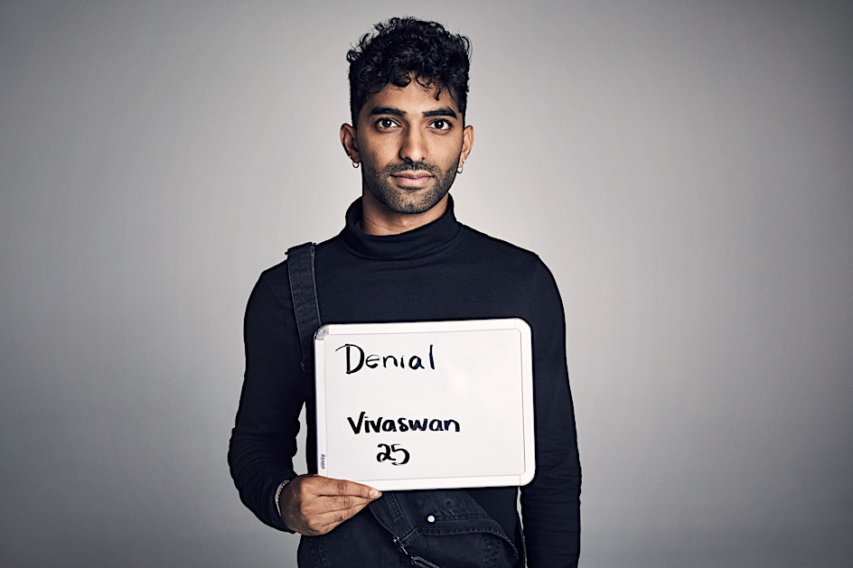 Young man with dark hair holds a whiteboard sign with the words 'Denial', 'Vivaswan', and '25' written in black marker indoors.