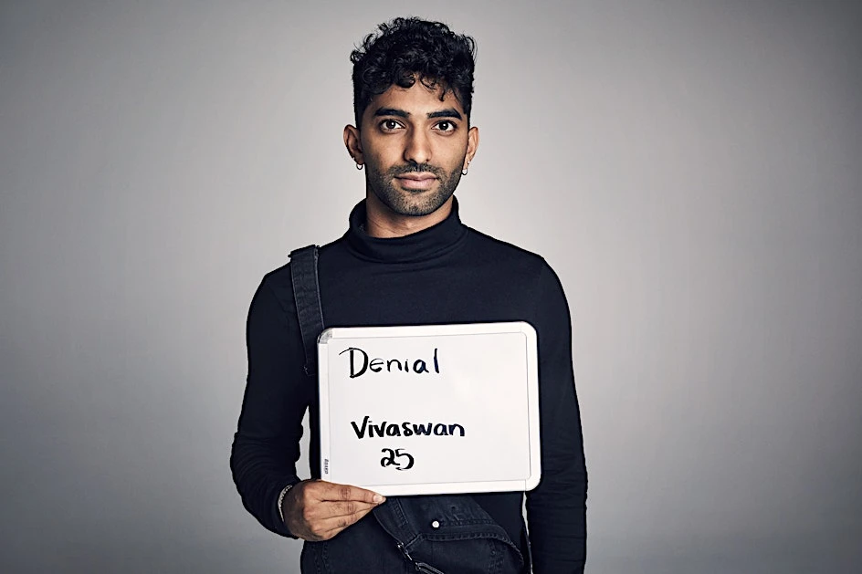 Young man with dark hair holds a whiteboard sign with the words 'Denial', 'Vivaswan', and '25' written in black marker indoors.