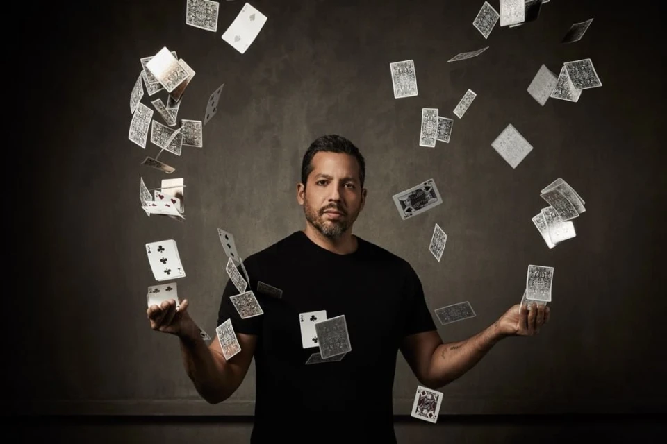 Adult male magician in black shirt performing card trick with deck of cards floating in air with dark background in a photo studio setting.