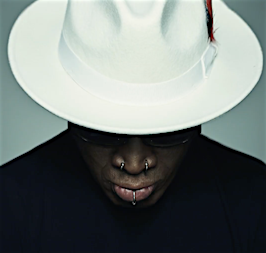 Close-up shot of a bald adult male wearing a white fedora with a red feather, a black turtleneck, sunglasses, and facial piercings.