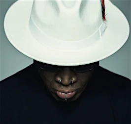 Close-up shot of a bald adult male wearing a white fedora with a red feather, a black turtleneck, sunglasses, and facial piercings.