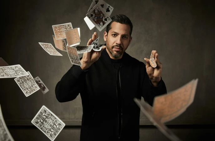 Magician performs card magic with playing cards, in studio environment, wearing a black jacket, and holding a playing card.
