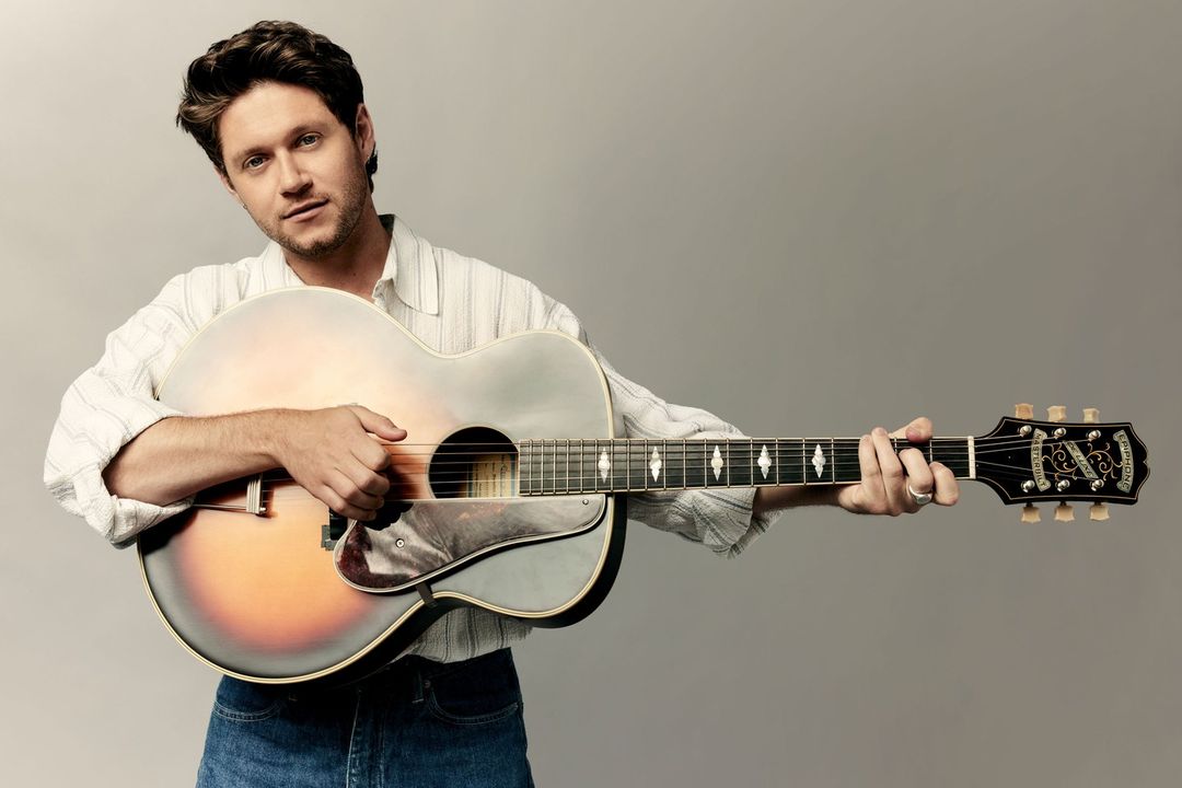 Adult male playing an acoustic guitar in a close-up studio shot; he wears a striped white shirt and blue jeans against a grey backdrop.