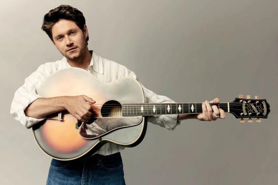 Adult male playing an acoustic guitar in a close-up studio shot; he wears a striped white shirt and blue jeans against a grey backdrop.