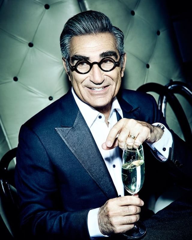 Portrait of an older man in a tuxedo and glasses smiling while holding a champagne flute against a quilted-leather background.