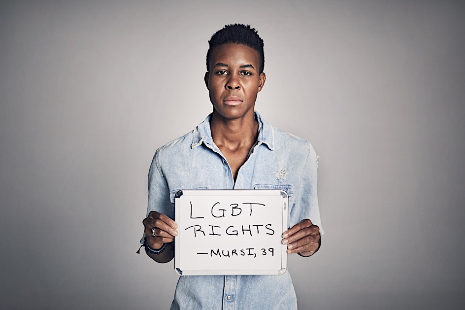 An adult Black person holds an LGBT Rights sign in a studio setting with soft lighting, wearing a denim shirt and neutral expression.