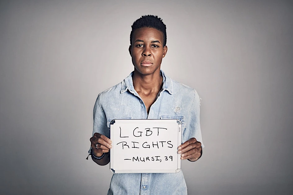 An adult Black person holds an LGBT Rights sign in a studio setting with soft lighting, wearing a denim shirt and neutral expression.