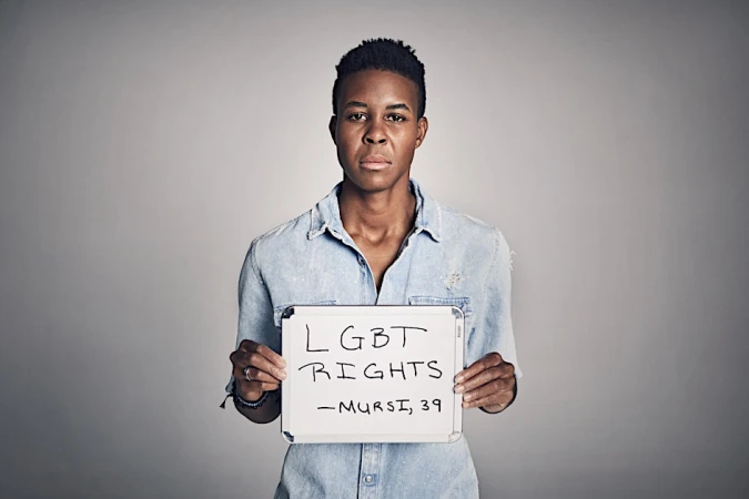 An adult Black person holds an LGBT Rights sign in a studio setting with soft lighting, wearing a denim shirt and neutral expression.