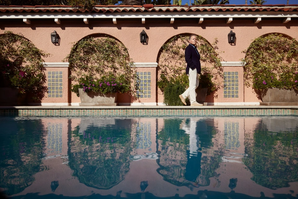 An adult male in a navy blazer walks alongside a pool with its water reflecting the facade of a salmon-colored building on a sunny day.