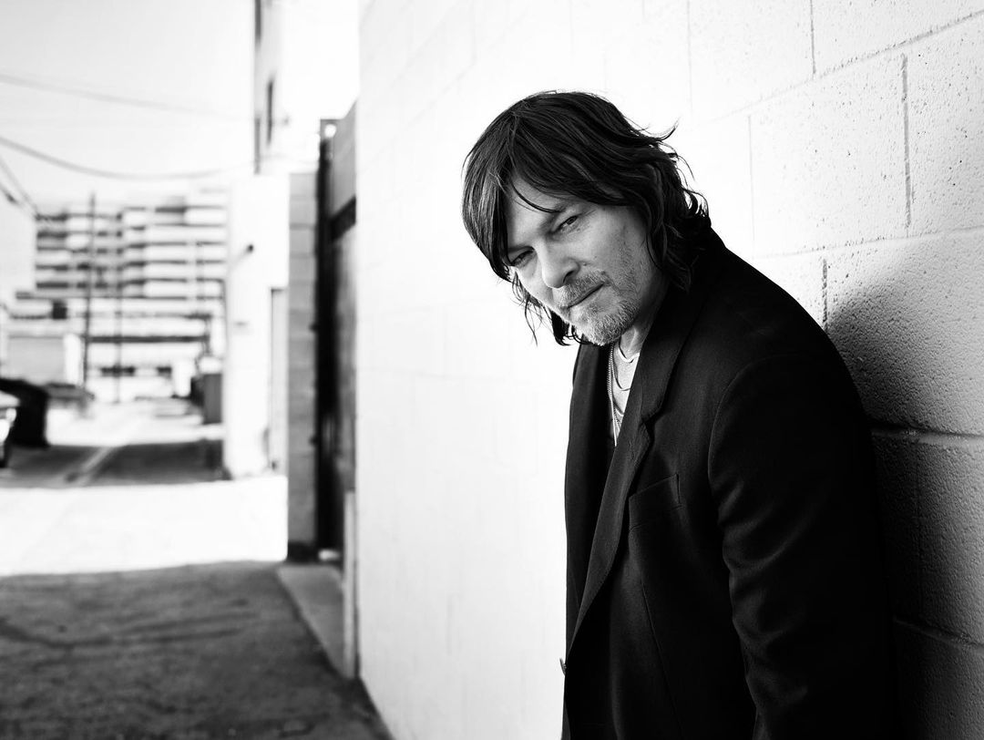 Black and white portrait of a man with shoulder-length hair, wearing a dark jacket and leaning against a brick wall in an urban setting.