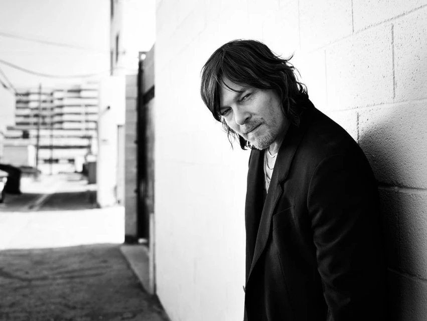 Black and white portrait of a man with shoulder-length hair, wearing a dark jacket and leaning against a brick wall in an urban setting.