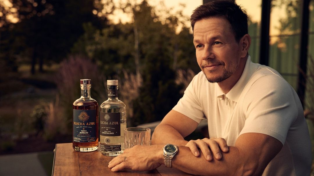Mark Wahlberg poses with Flecha Azul tequila bottles on a wooden table outdoors with an out-of-focus green background.