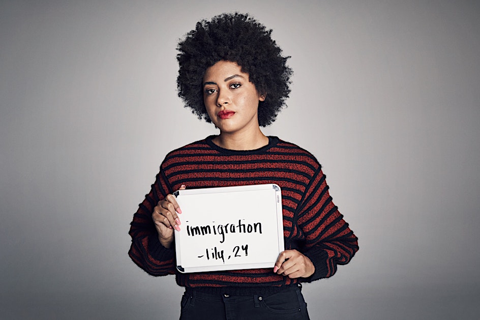 Portrait of a woman with an afro holding a sign with text. She wears a striped sweater, posing against a solid grey backdrop in studio lighting.