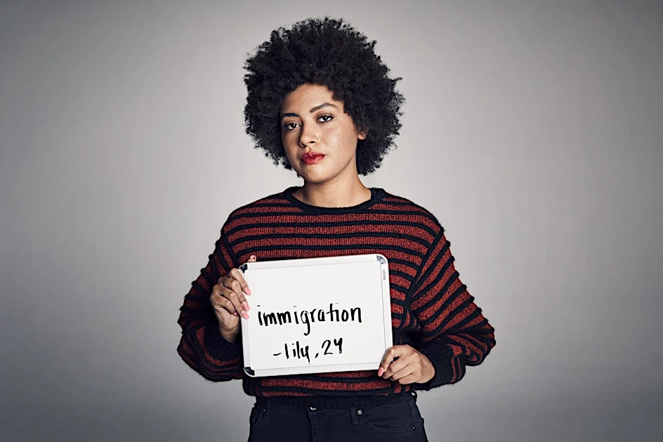 Portrait of a woman with an afro holding a sign with text. She wears a striped sweater, posing against a solid grey backdrop in studio lighting.