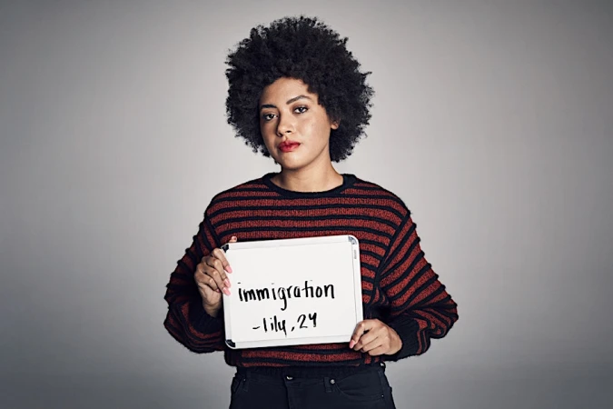 Portrait of a woman with an afro holding a sign with text. She wears a striped sweater, posing against a solid grey backdrop in studio lighting.