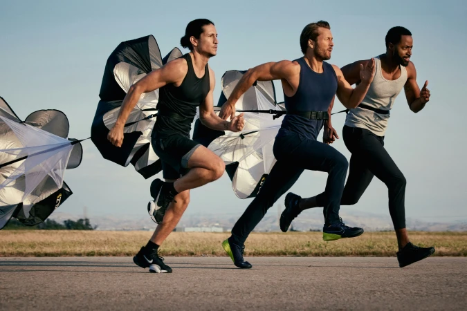 Three athletic men run outdoors, each pulling a black and white parachute for resistance training on a paved surface.
