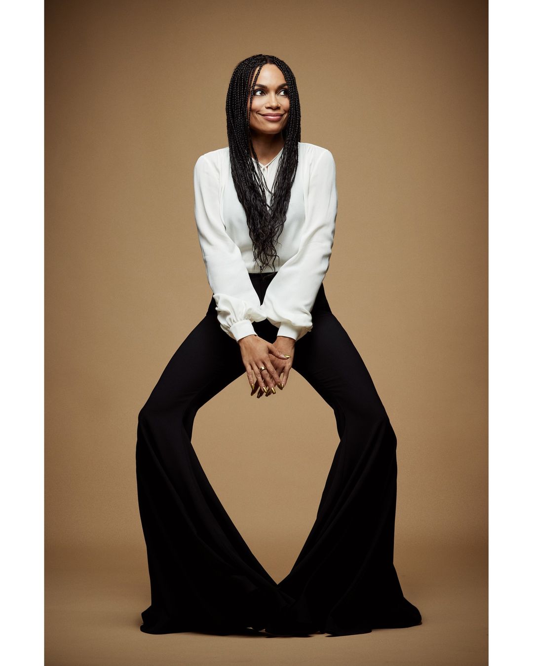 Full-length portrait of a woman with long dark braids, wearing a white blouse and black bell-bottoms, posing in a photo studio with a tan backdrop.