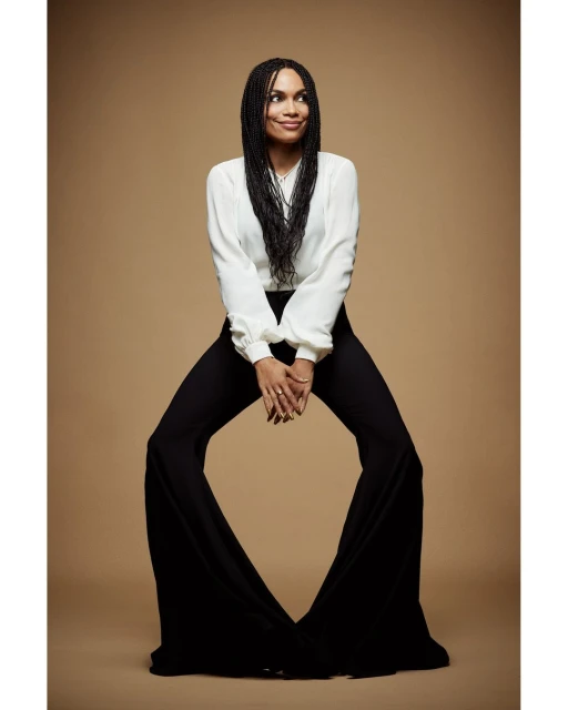 Full-length portrait of a woman with long dark braids, wearing a white blouse and black bell-bottoms, posing in a photo studio with a tan backdrop.