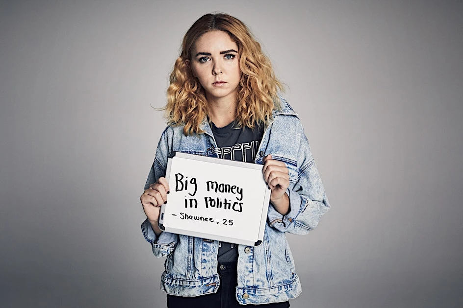 Young woman with curly hair and a denim jacket holds a white sign with the text 'Big Money in Politics - Shawnee, 25'.