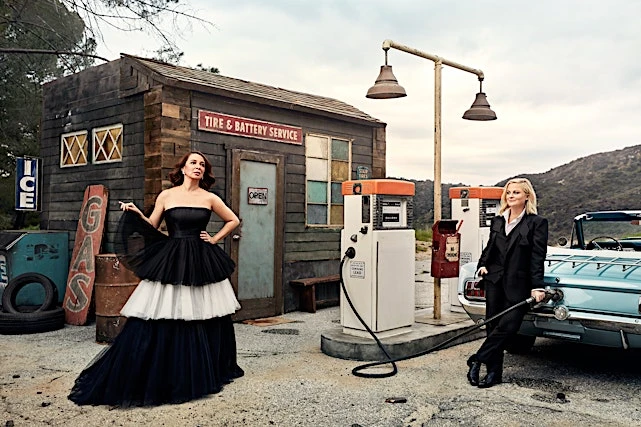 Tina Fey and Amy Poehler pose in front of a gas station, one in a gown and the other in a suit, with a vintage car nearby.