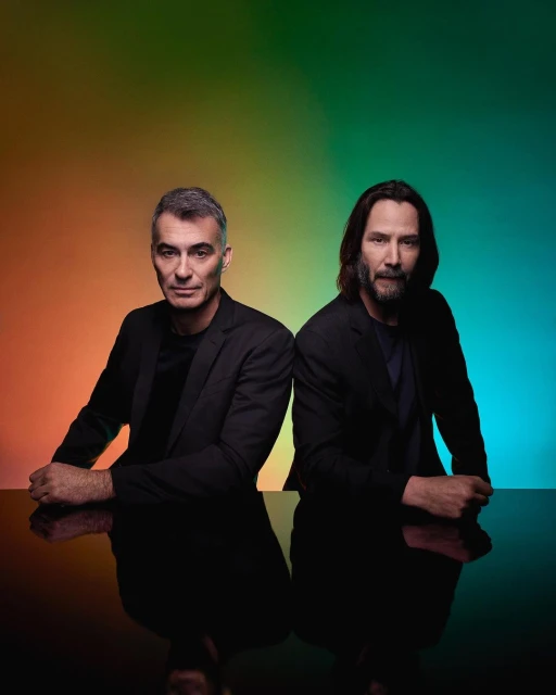 Portrait of two adult males in dark suits, posing in front of a gradient orange, green, and blue background with reflective surface.