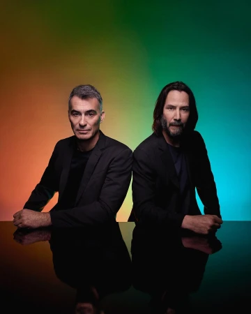 Portrait of two adult males in dark suits, posing in front of a gradient orange, green, and blue background with reflective surface.