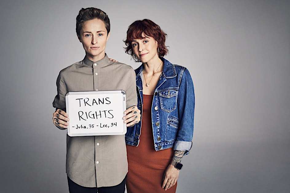 Two adults holding a sign advocating for transgender rights, standing against a neutral grey background in a studio setting.