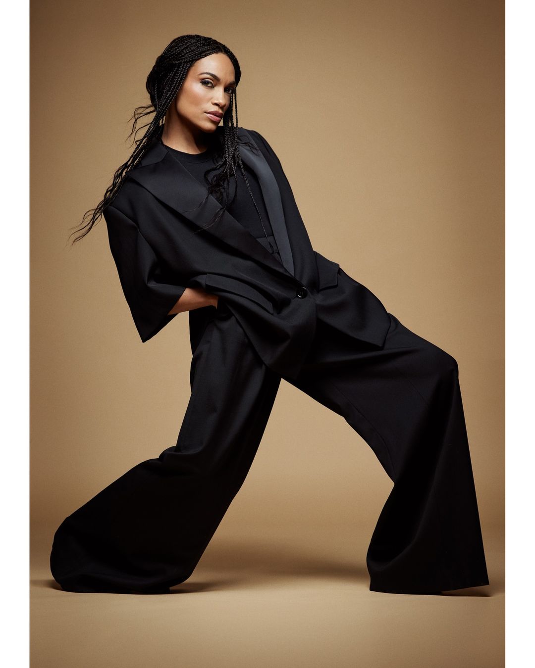 Model with long braided hair wearing an oversized black suit posing against a warm neutral background