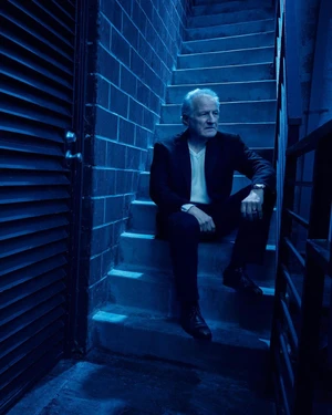 An older man in a dark suit sitting on concrete outdoor stairs at night under cool blue lighting.