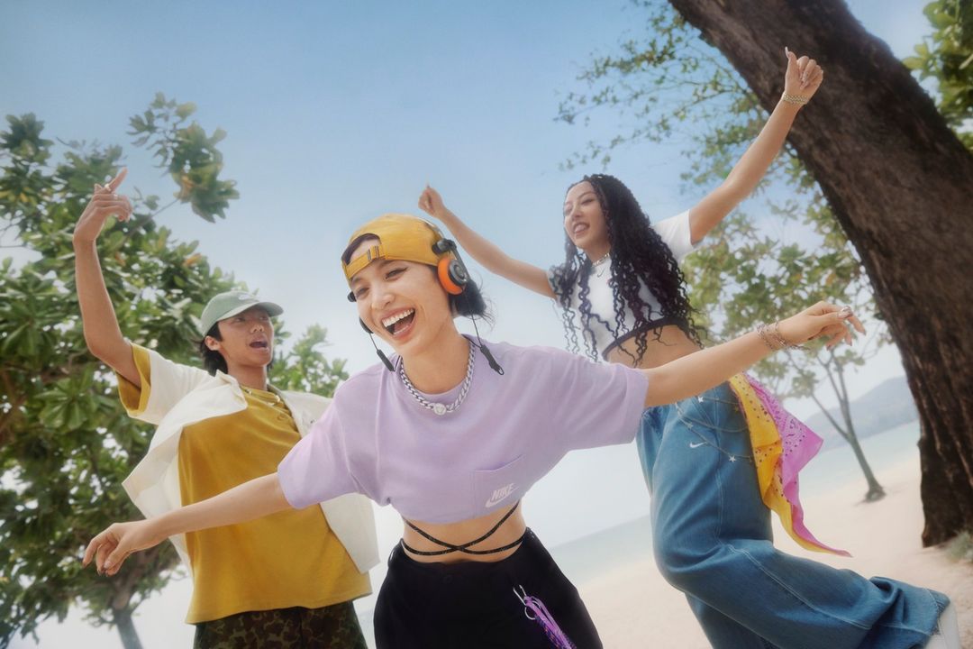 Three young adults joyfully celebrate on a sunny beach, wearing trendy casual clothing and Nike branded apparel, set against a blue sky.