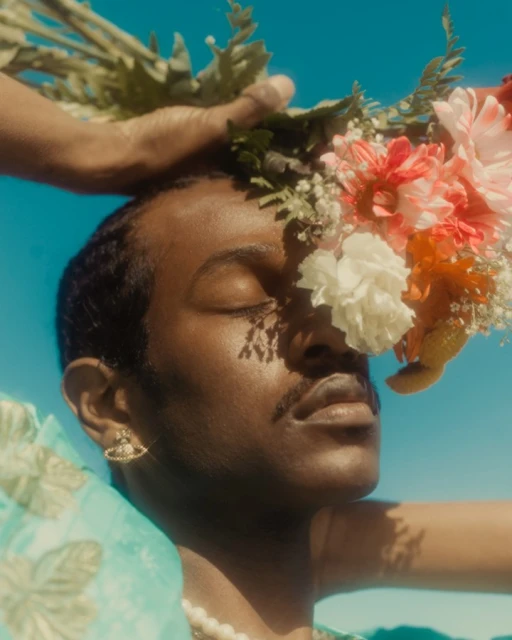 Portrait of a Black man with eyes closed, adorned with flowers, set against a bright blue sky, featuring an angelic, serene aesthetic.